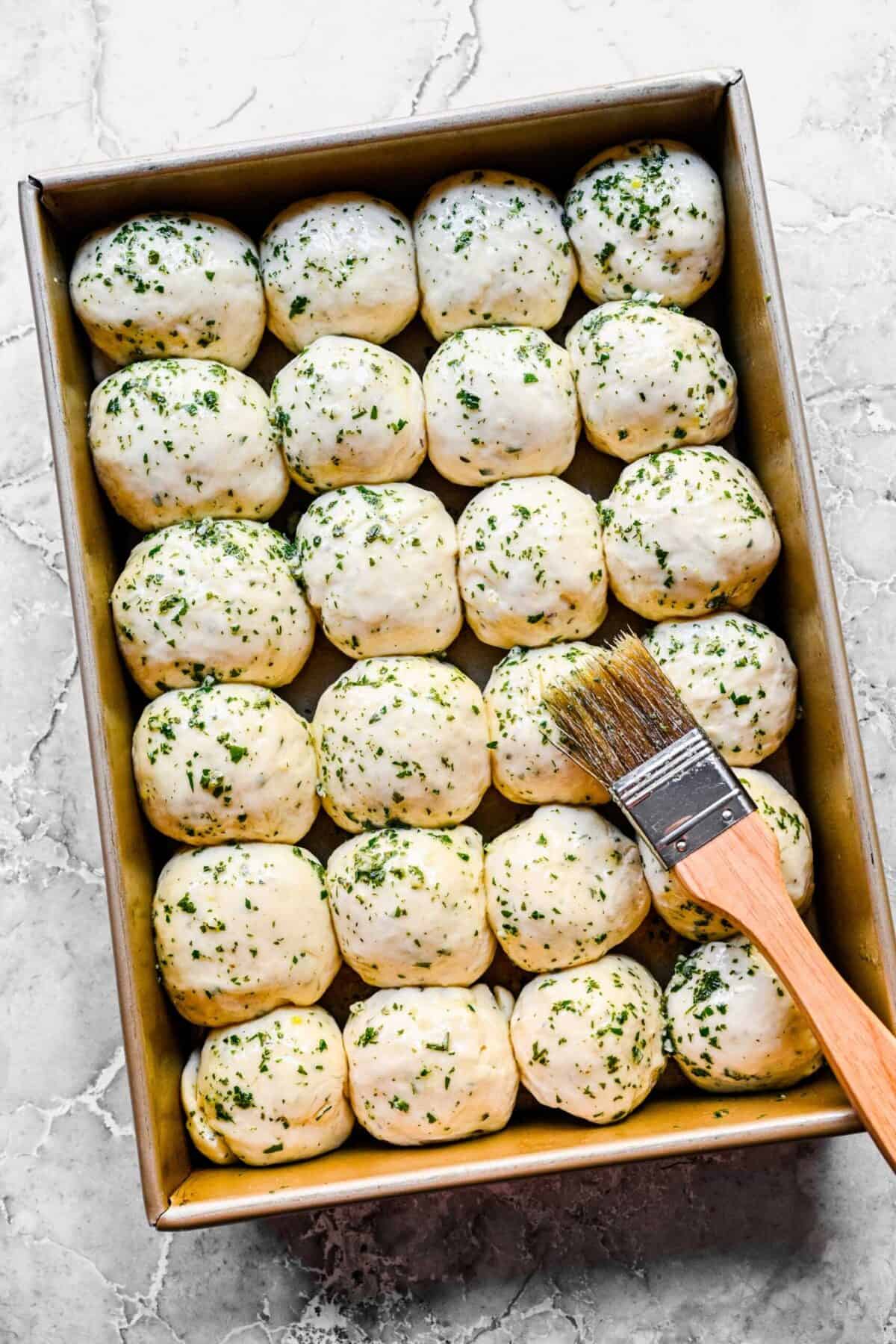 A baking tray filled with 24 balls of uncooked garlic herb roll dough, covered in a butter, garlic, and herb mixture, with a baking brush on top