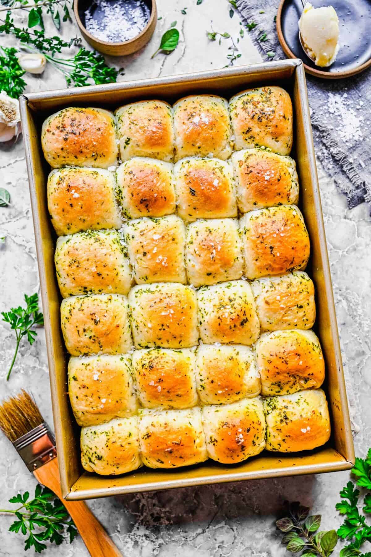 A tray of 24 garlic herb parker house rolls, fully baked, topped with herbs, next to some fresh herbs and a baking brush