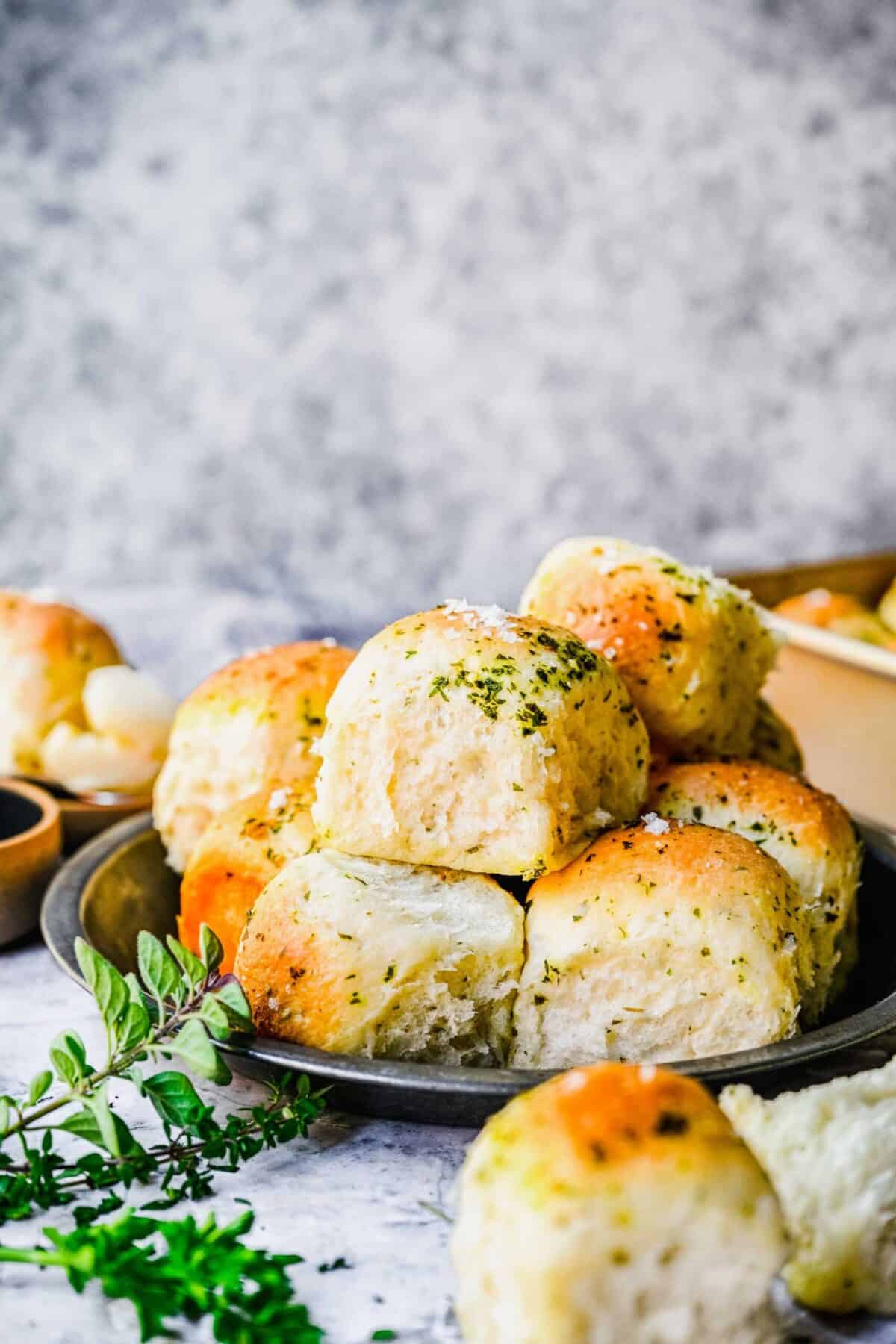 A plate with a pile of garlic herb parker house rolls, next to some fresh herbs, with more rolls in the foreground and background.