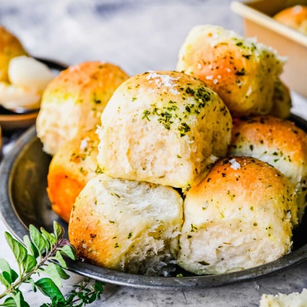 Close up of a plate with garlic herb parker house rolls, topped with garlic and herbs, next to some fresh herbs.