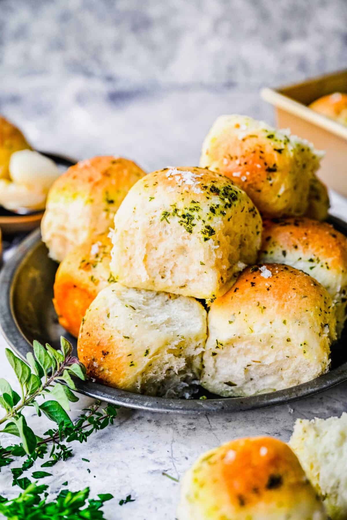 Close up of a plate with garlic herb parker house rolls, topped with garlic and herbs, next to some fresh herbs.
