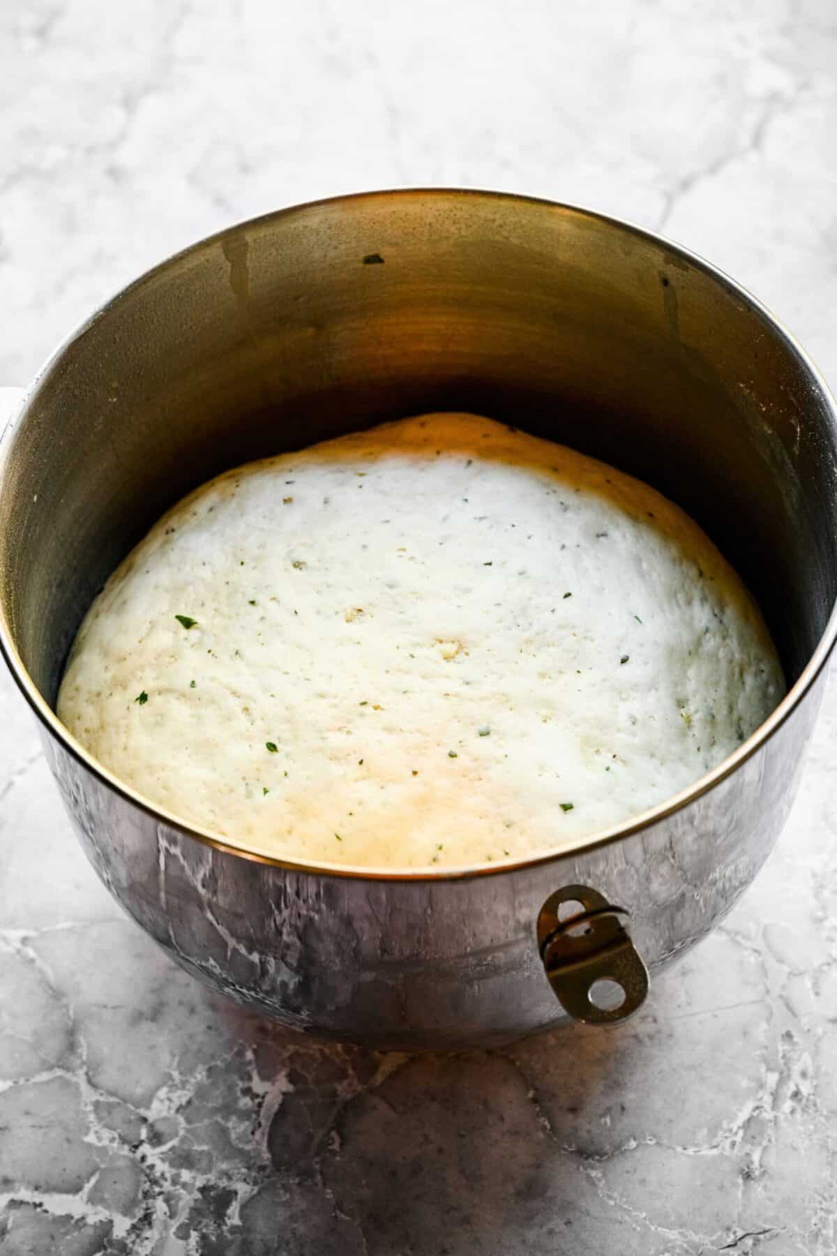 A stand mixer filled with puffy garlic herb roll dough that has risen.