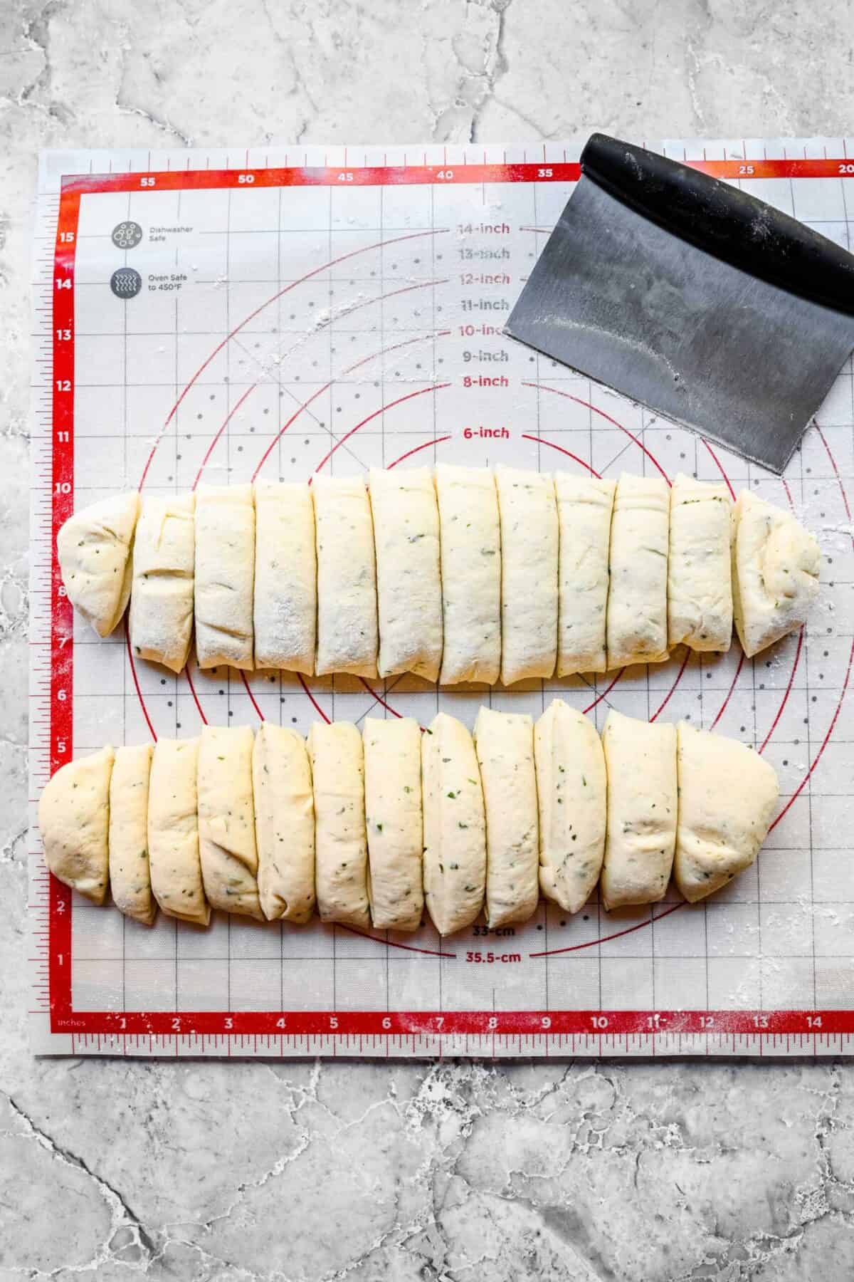 Two logs of garlic herb parker house roll dough, each cut into 12 pieces, on top of a silicone mat with measurements, next to a bench scraper.