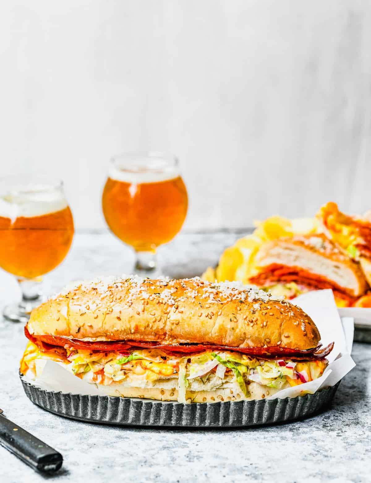 A large grinder sandwich on a tray, with two glasses of beer and a bag of chips in the background