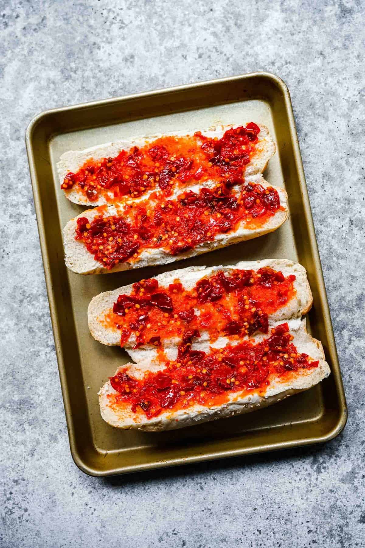 Two hoagie loaves on a baking sheet, covered in Calabrian chili paste