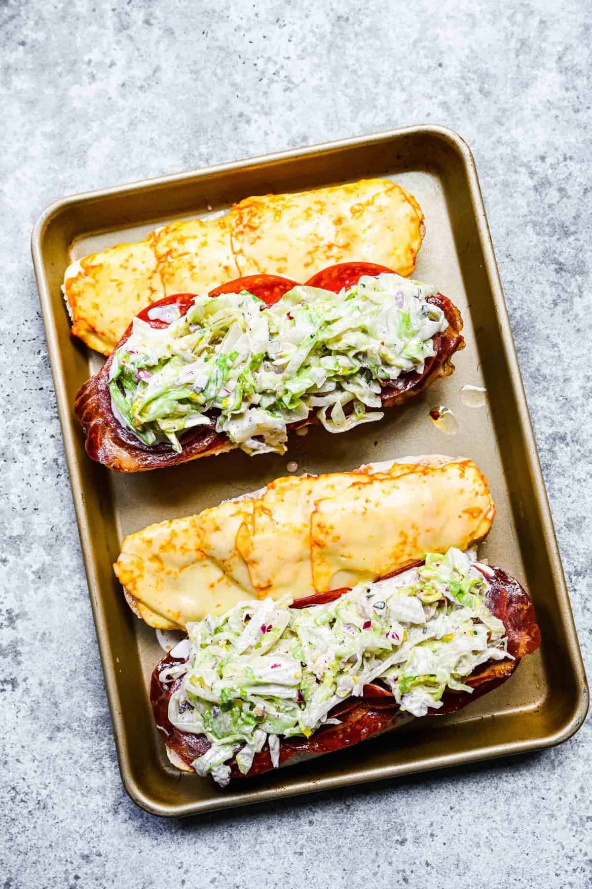 Two hoagie rolls on a baking sheet, with melted cheese on one half, and cured meats topped with a salad on the other half