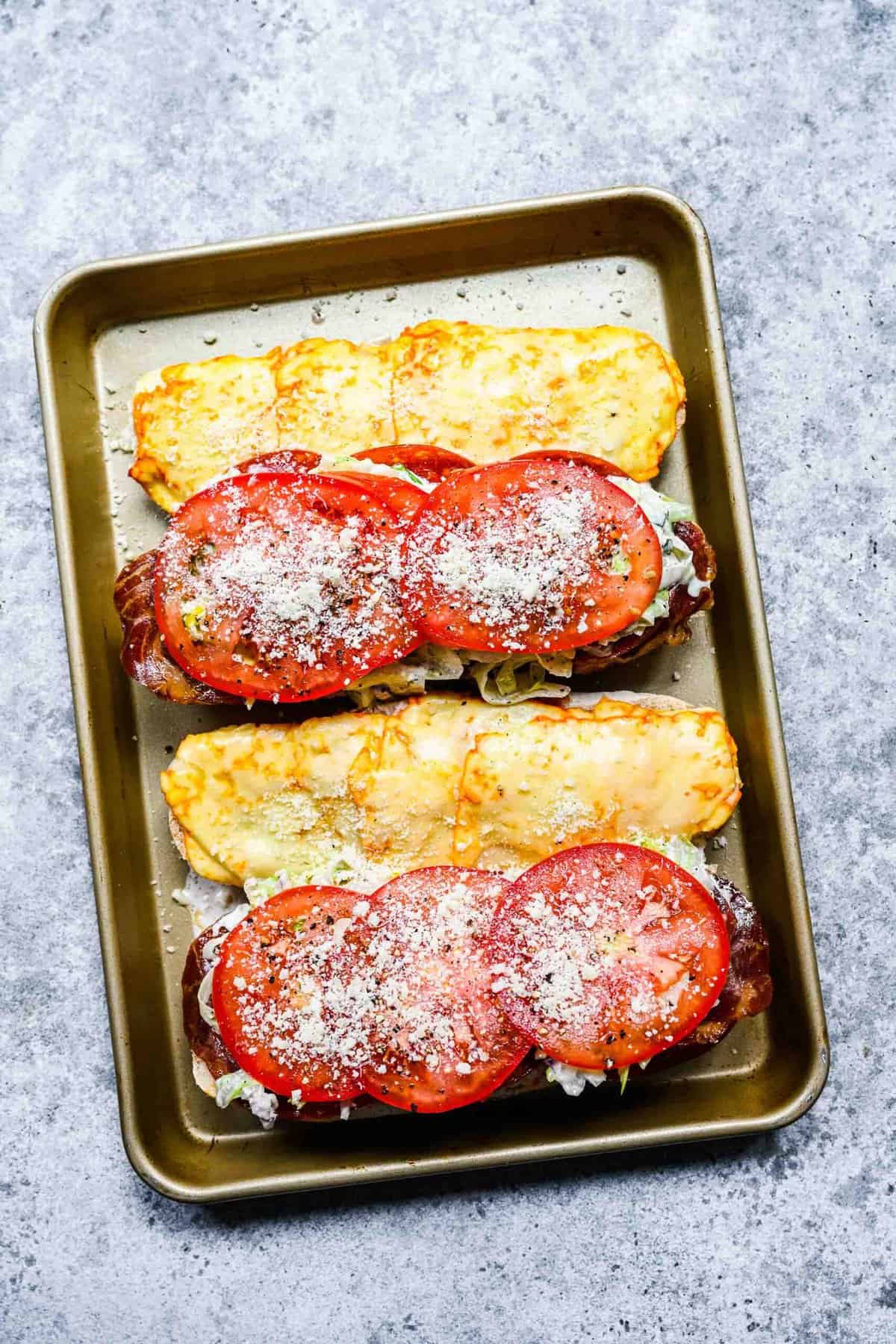 Two hoagie loaves on a baking sheet, with melted cheese on one half, and cured meats, topped with a salad, topped with slices of tomato with salt, pepper, and parmesan on top on the other half