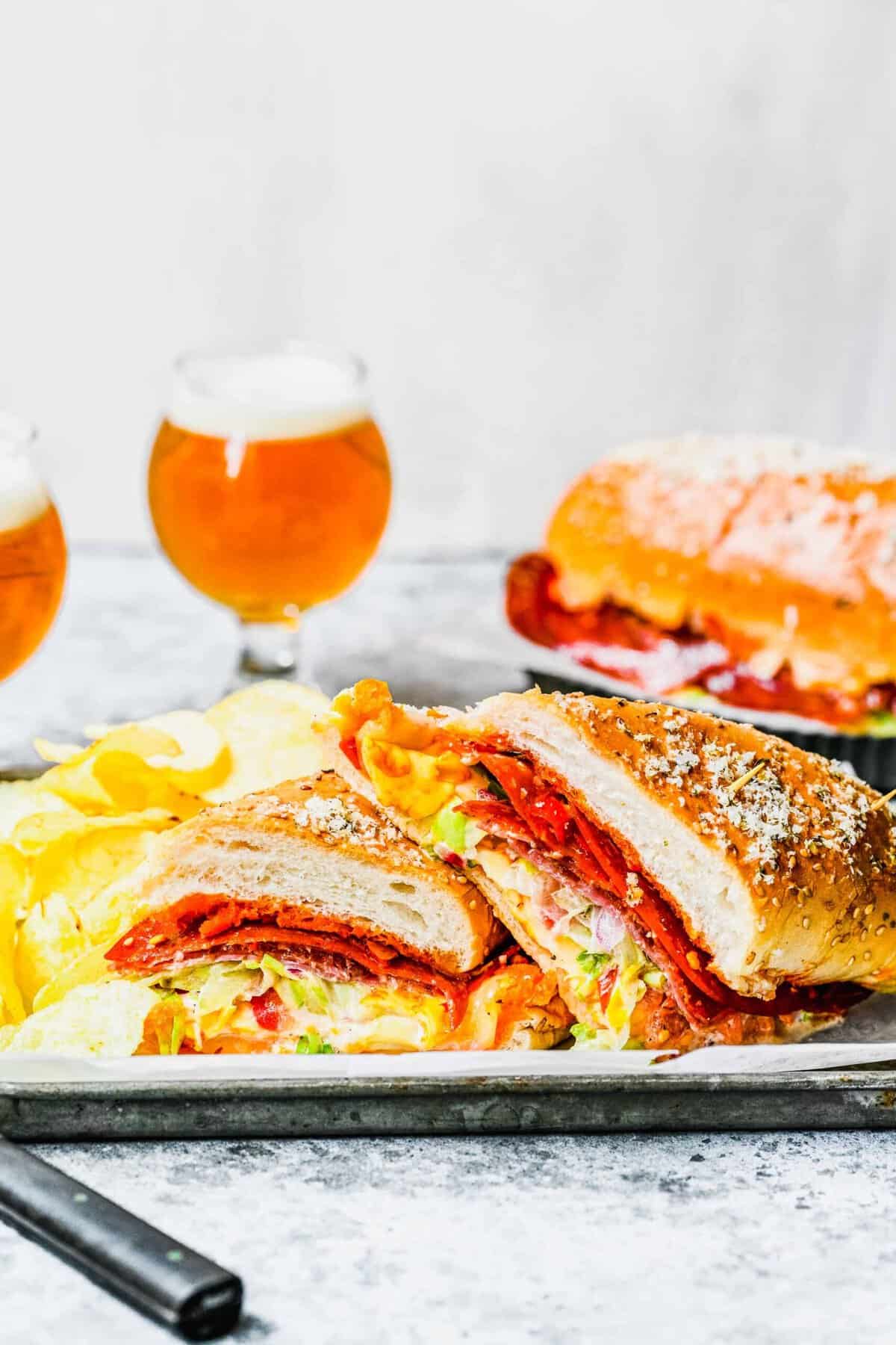 A plate with a grinder sandwich cut in half, with two glasses of beer and a second sandwich in the background