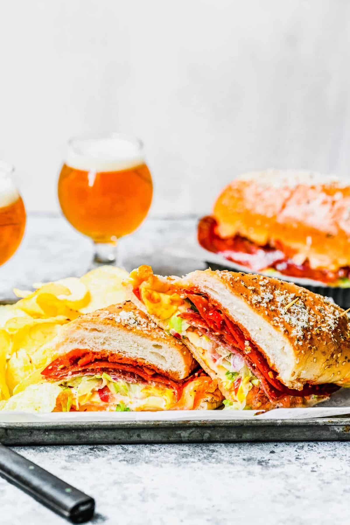 This grinder sandwich is packed with three meats, three cheeses, and spice, fresh, acidic vegetables, making it the ultimate sandwich. A plate with a grinder sandwich cut in half, with two glasses of beer and a second sandwich in the background