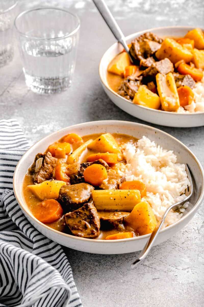 beef, potatoes, celery, and carrots in a stew-like sauce on a bed of white rice in a rimmed ceramic bowl with a fork next to it. a striped linen towel and a glass of water can be seen