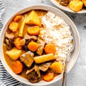 overhead image of beef, potatoes, carrots, celery in a stew-like sauce on a bed of white rice with a fork in a rimmed ceramic bowl next to another bowl of the same contents.