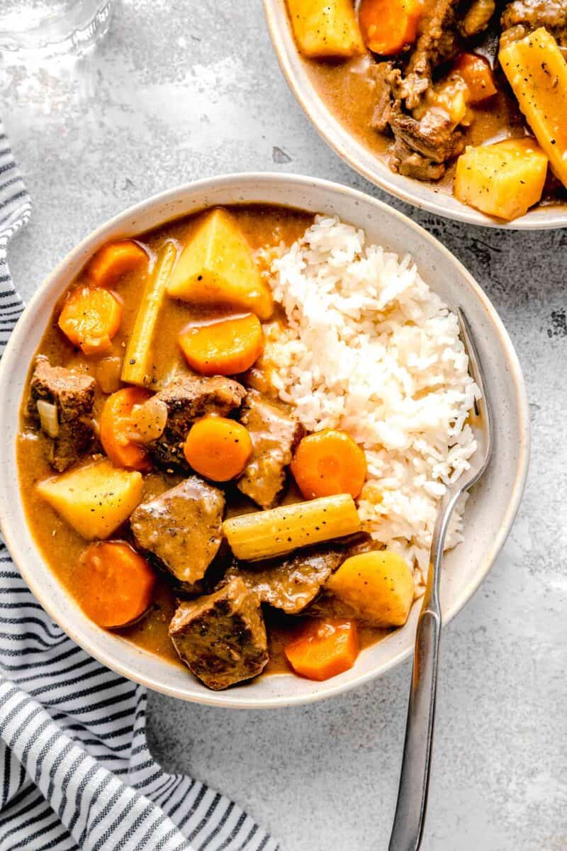 overhead image of beef, potatoes, carrots, celery in a stew-like sauce on a bed of white rice with a fork in a rimmed ceramic bowl next to another bowl of the same contents.