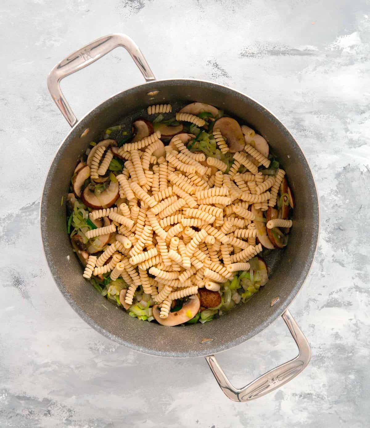 uncooked spiral pasta being added to pot with cooked leeks, onions, garlic, and mushrooms