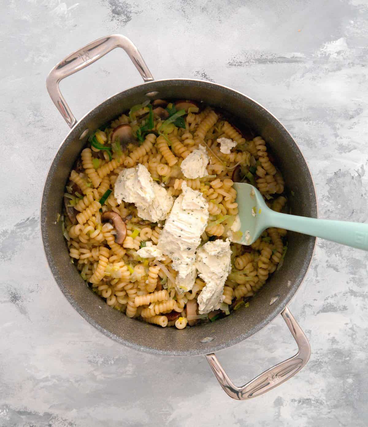 boursin cheese being added to cooked pasta, leeks, onions, garlic, and mushrooms