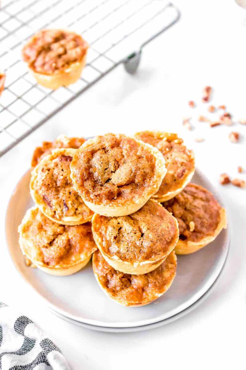 pecan tassies stacked on two white plates next to a wire baking rack with more pecan tassies on top
