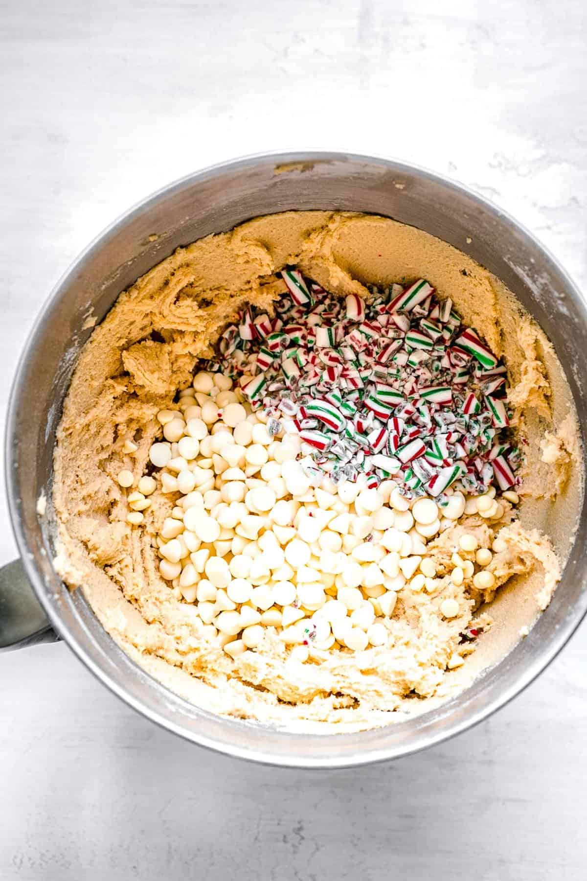 white chocolate chips and crushed candy canes sitting on top of the cookie dough in a metal bowl