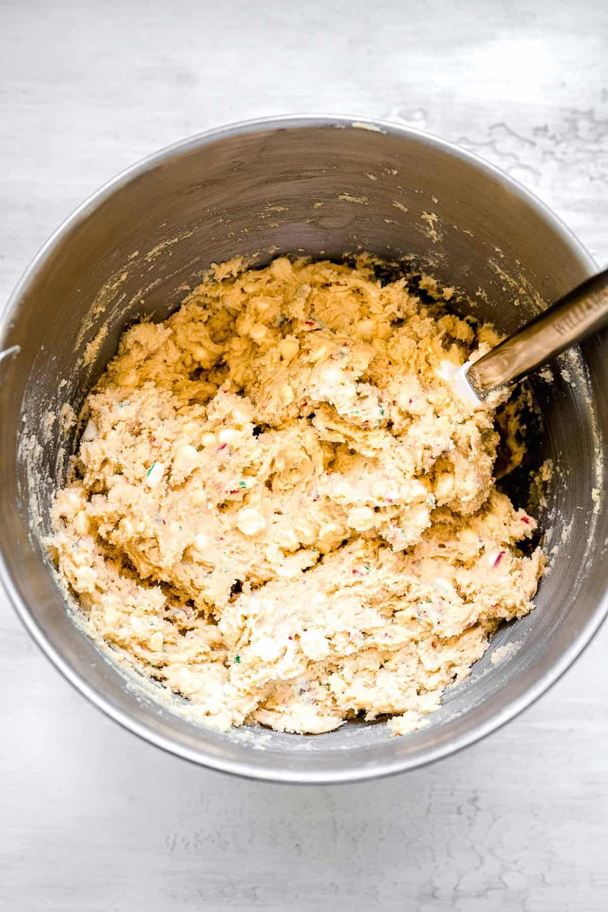 white chocolate chips and crushed candy canes being mixed with the dough with a spatula in a metal bowl
