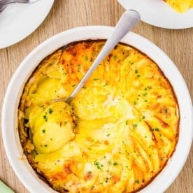 overhead image of a wooden cutting board and potatoes au gratin in a white casserole dish with a silver spoon that has scooped out some of the side dish.