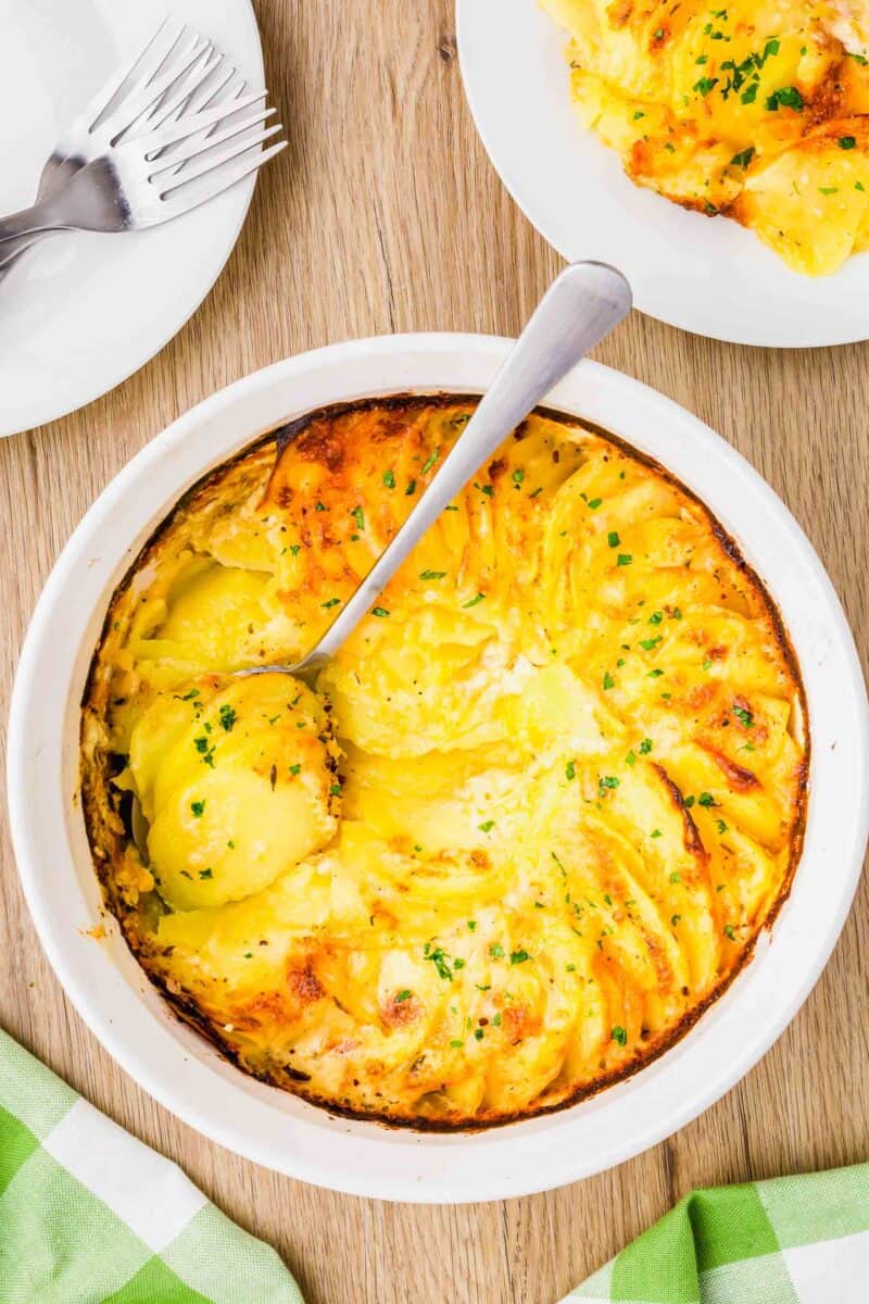 overhead image of a wooden cutting board and potatoes au gratin in a white casserole dish with a silver spoon that has scooped out some of the side dish.