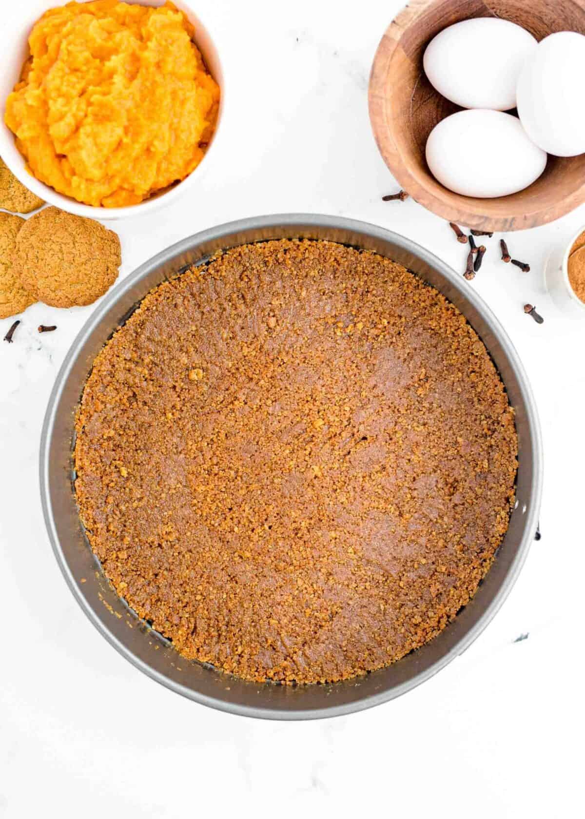 Gingersnap crust in a springform pan, surrounded by gingersnaps, eggs, and a bowl of pumpkin puree