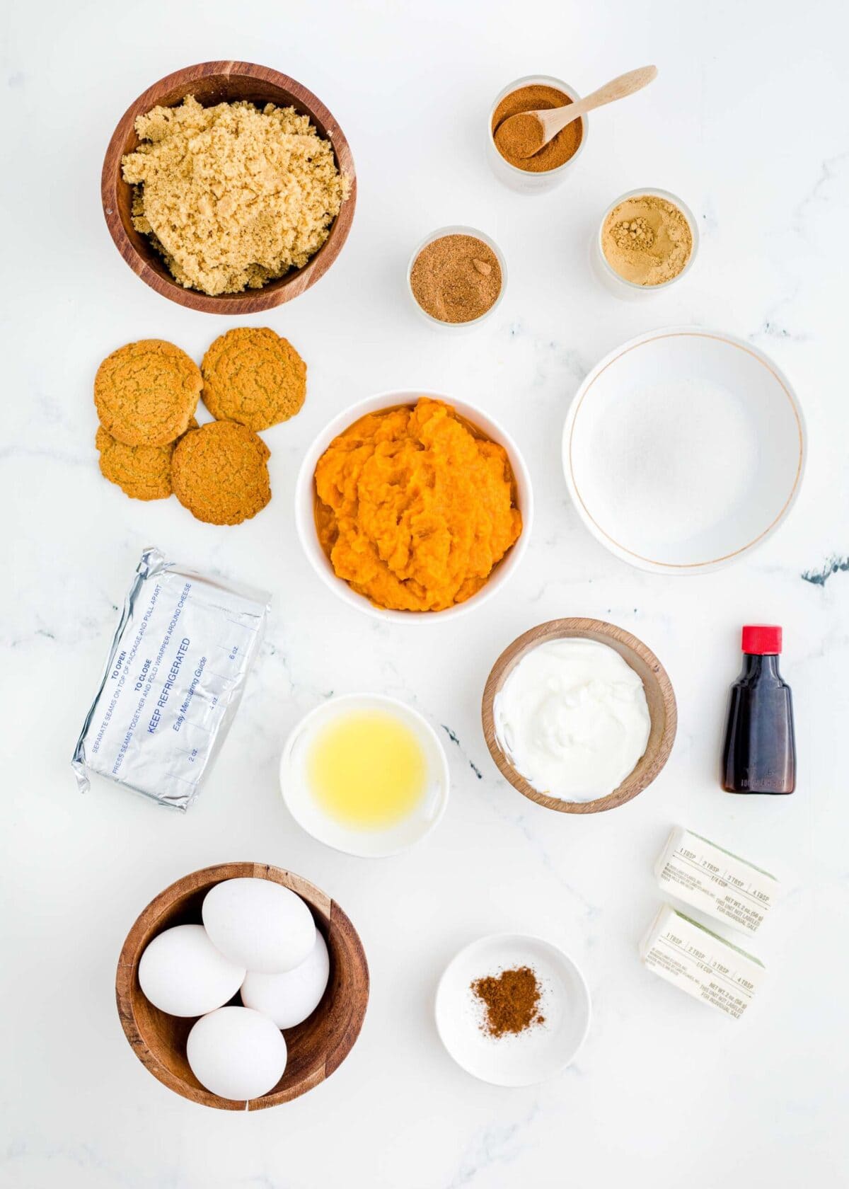 Overhead view of the ingredients for pumpkin cheesecake: a bowl of brown sugar, a bowl of white sugar, a bowl of pumpkin puree, a bowl of melted butter, a bowl of eggs, four bowls of spices, a bowl of sour cream, a package of cream cheese, two sticks of butter, a jar of vanilla extract, and some gingersnap cookies