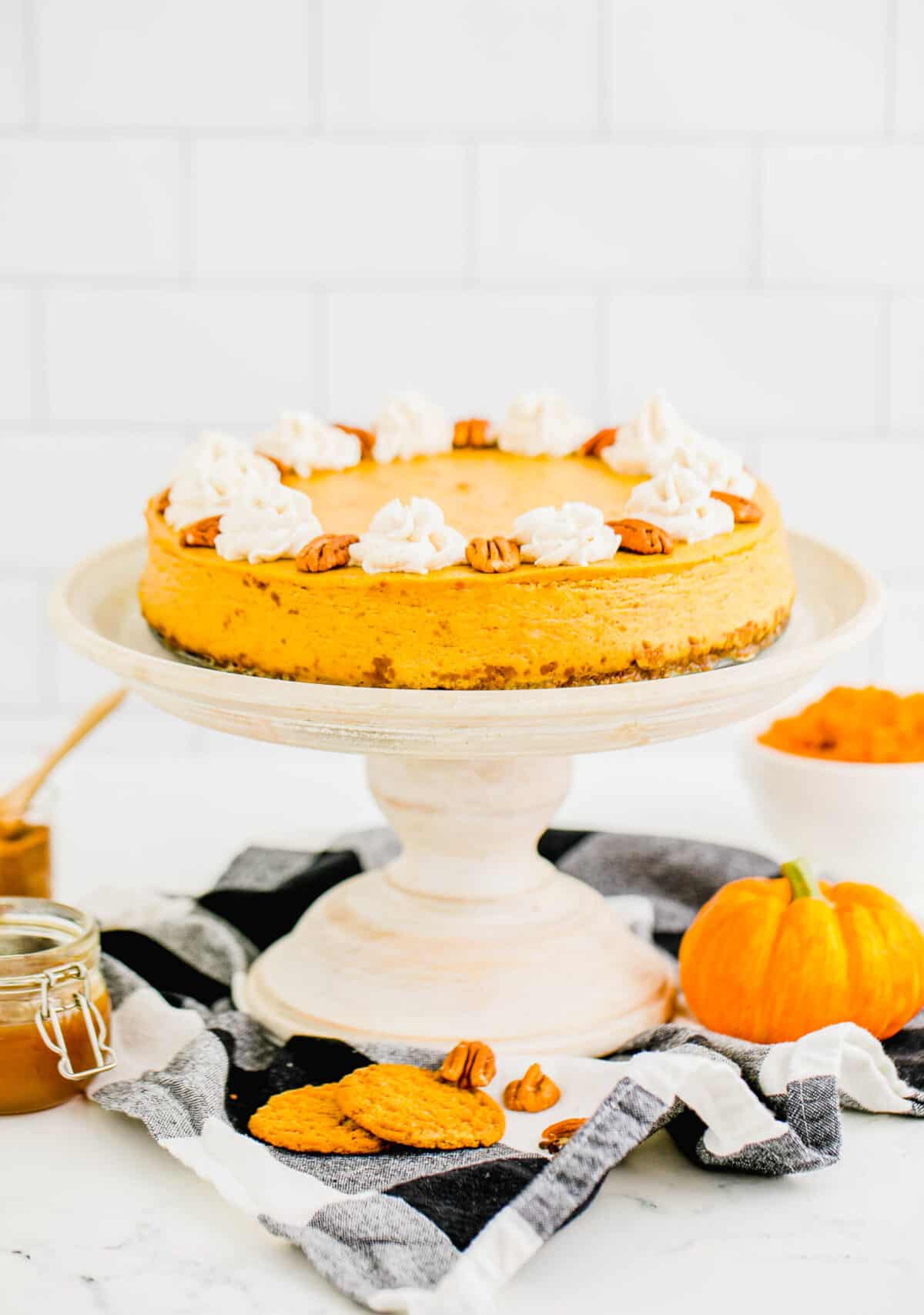 A pumpkin cheesecake on a cake stand, topped with pecans and whipped cream, surrounded by mini pumpkins