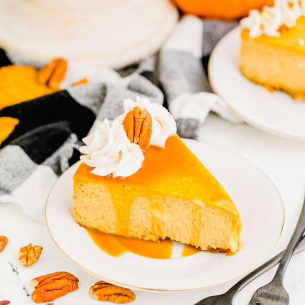 A slice of pumpkin cheesecake on a plate, topped with whipped cream, a pecan, and caramel sauce, with two forks next to it