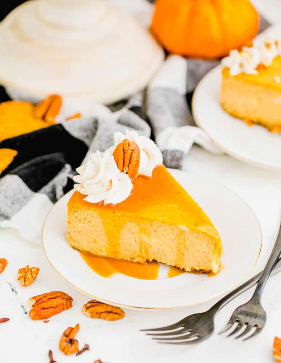 A slice of pumpkin cheesecake on a plate, topped with whipped cream, a pecan, and caramel sauce, with two forks next to it