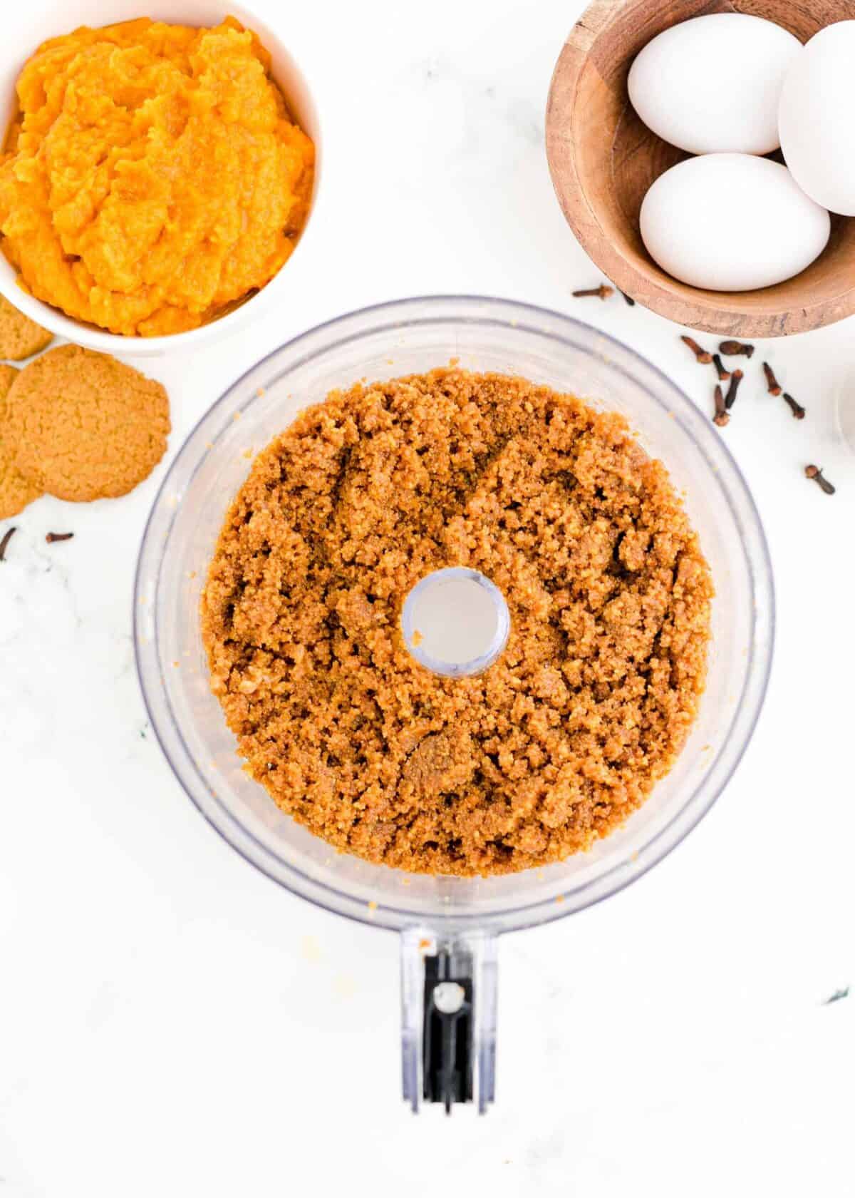 A food processor with pulsed gingersnap cookies and butter, surrounded by cookies, eggs, and a bowl of pumpkin puree