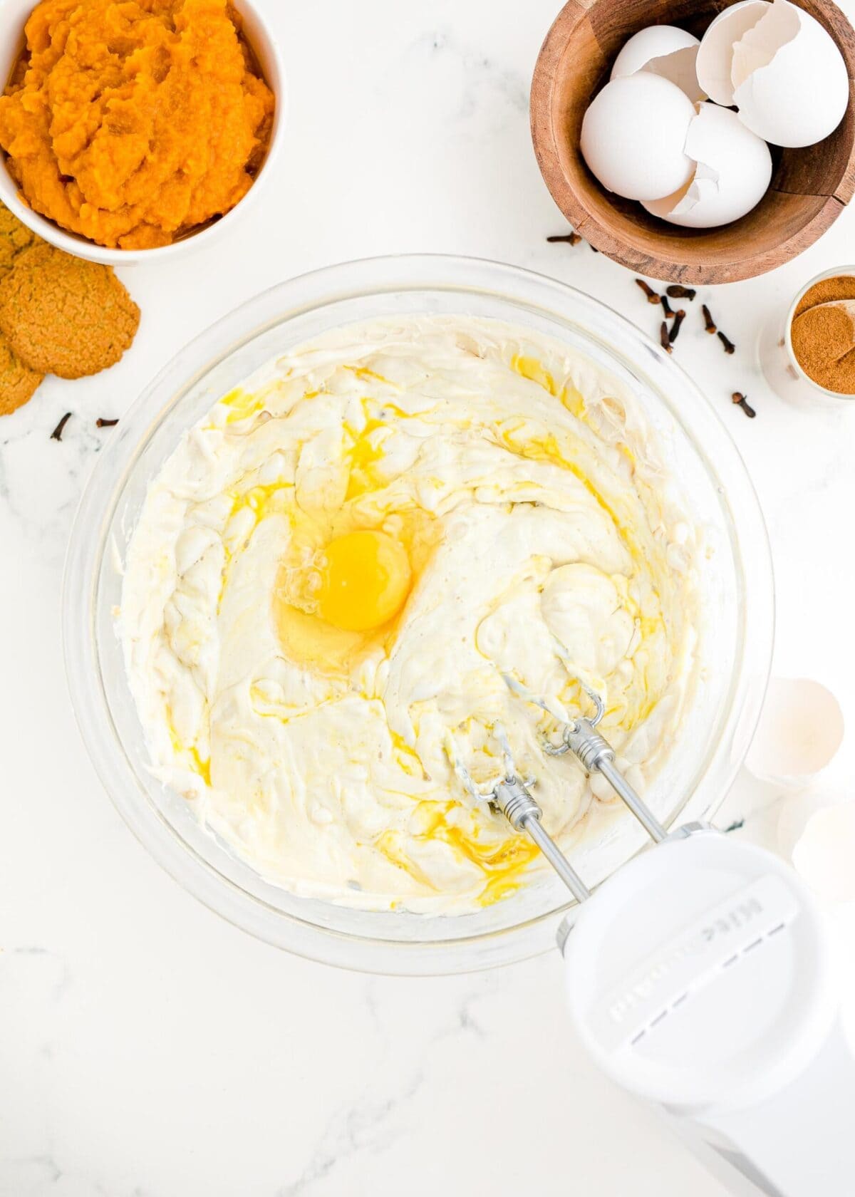 A mixing bowl with blended cream cheese, brown sugar, and white sugar in it, with an egg on top and an electric mixer in the bowl