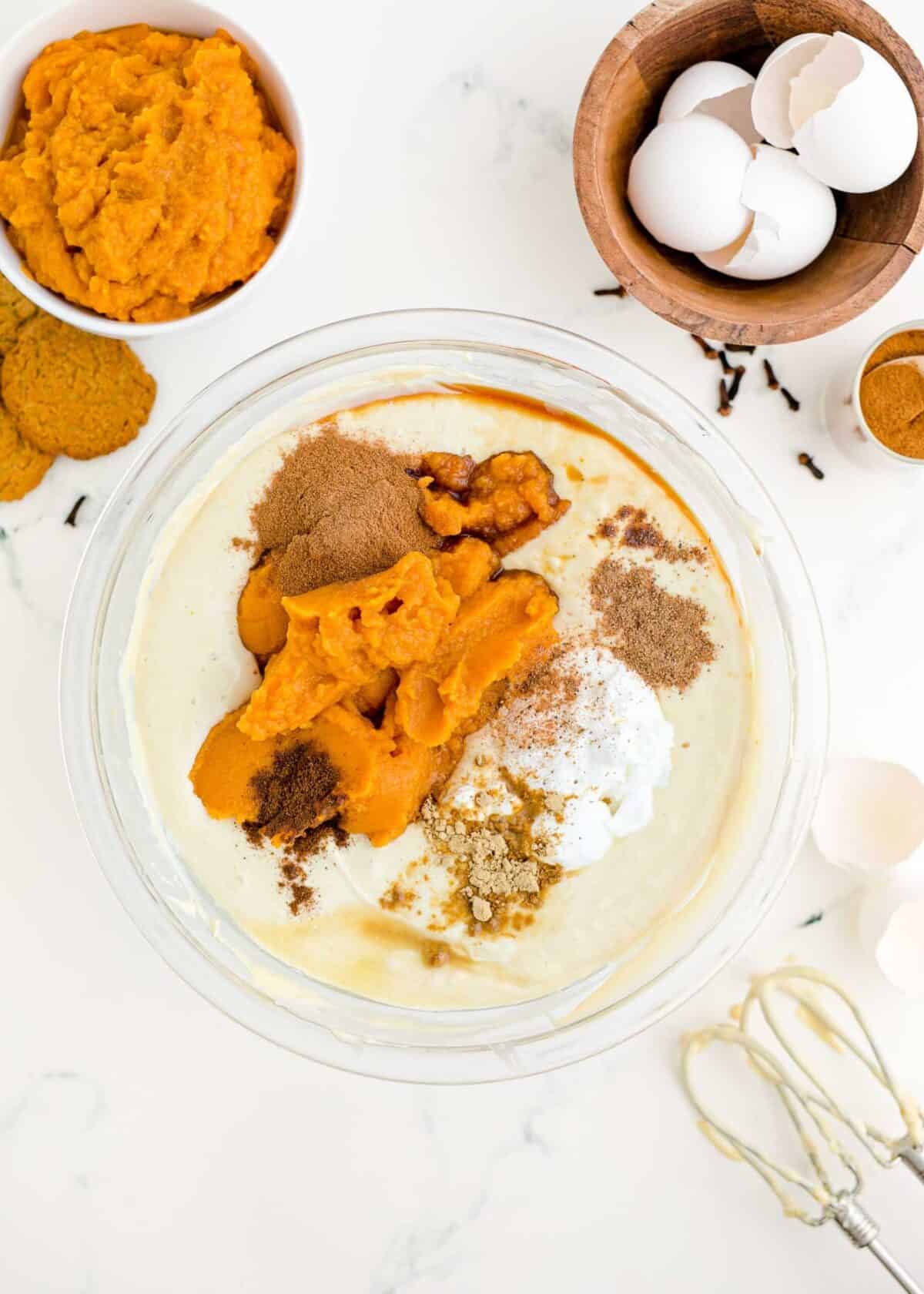 A mixing bowl filled with a cream cheese, sugar, and egg mixture, topped with unmixed pumpkin puree, sour cream, vanilla, lemon juice, and spices