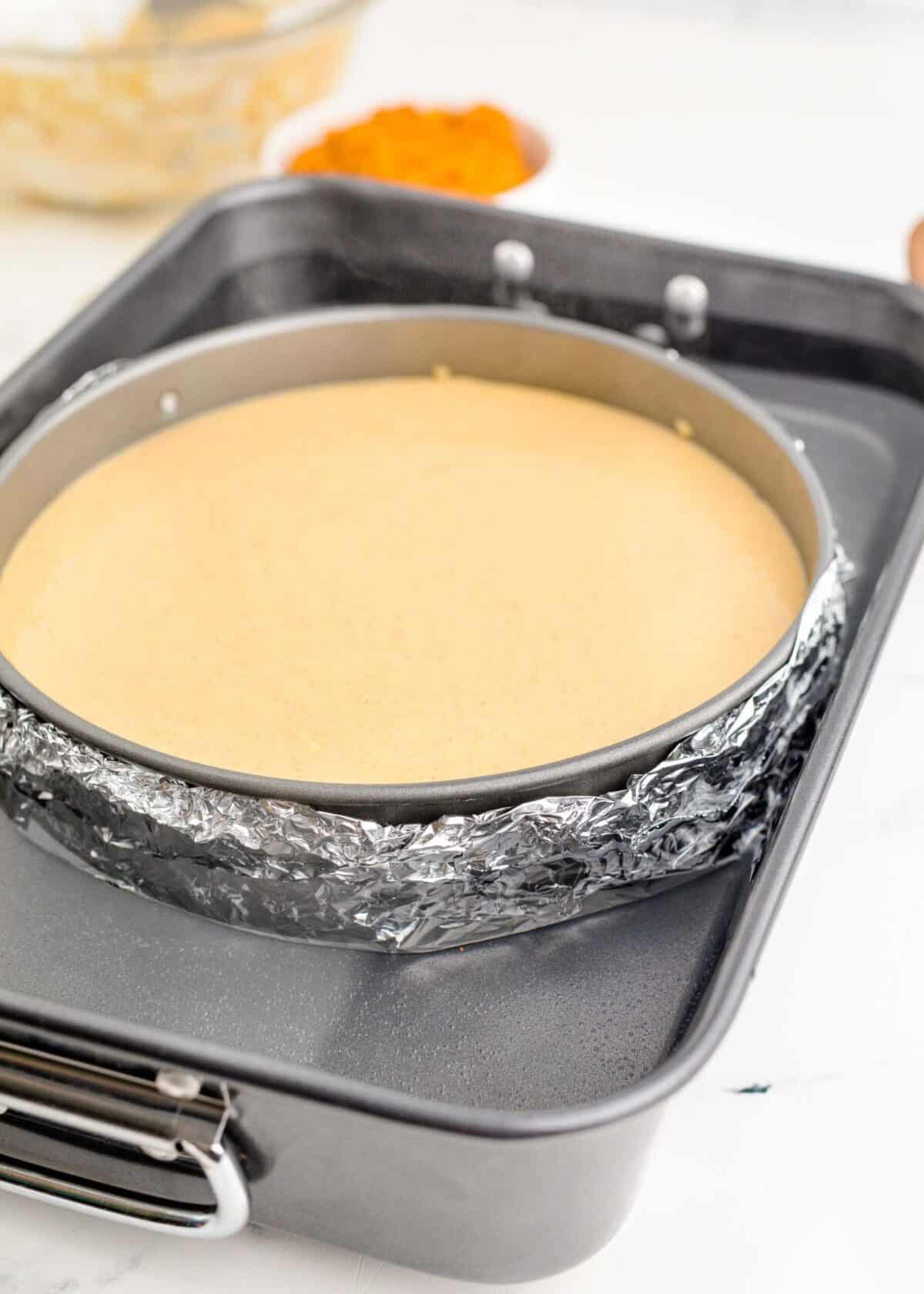 A springform pan filled with pumpkin cheesecake batter, wrapped in aluminum foil, in a water bath