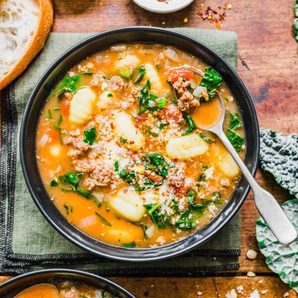 A bowl of sausage gnocchi soup with a spoon in it, next to another bowl of soup, some leaves of kale, and a small bowl of chili flakes