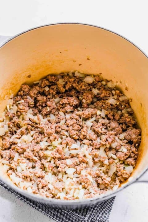 A pot with cooked, broken up sausage in it, as well as onions