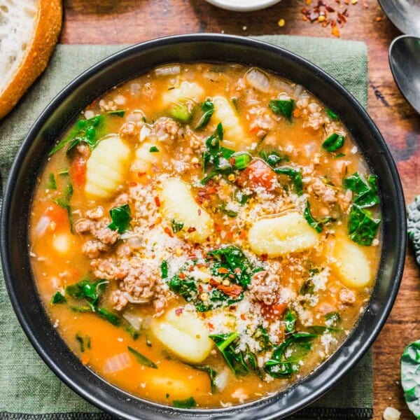 A bowl of sausage gnocchi soup on top of a kitchen towel, next to a bowl of parmesan cheese, a bowl of chili flakes, some bread, and some spoons