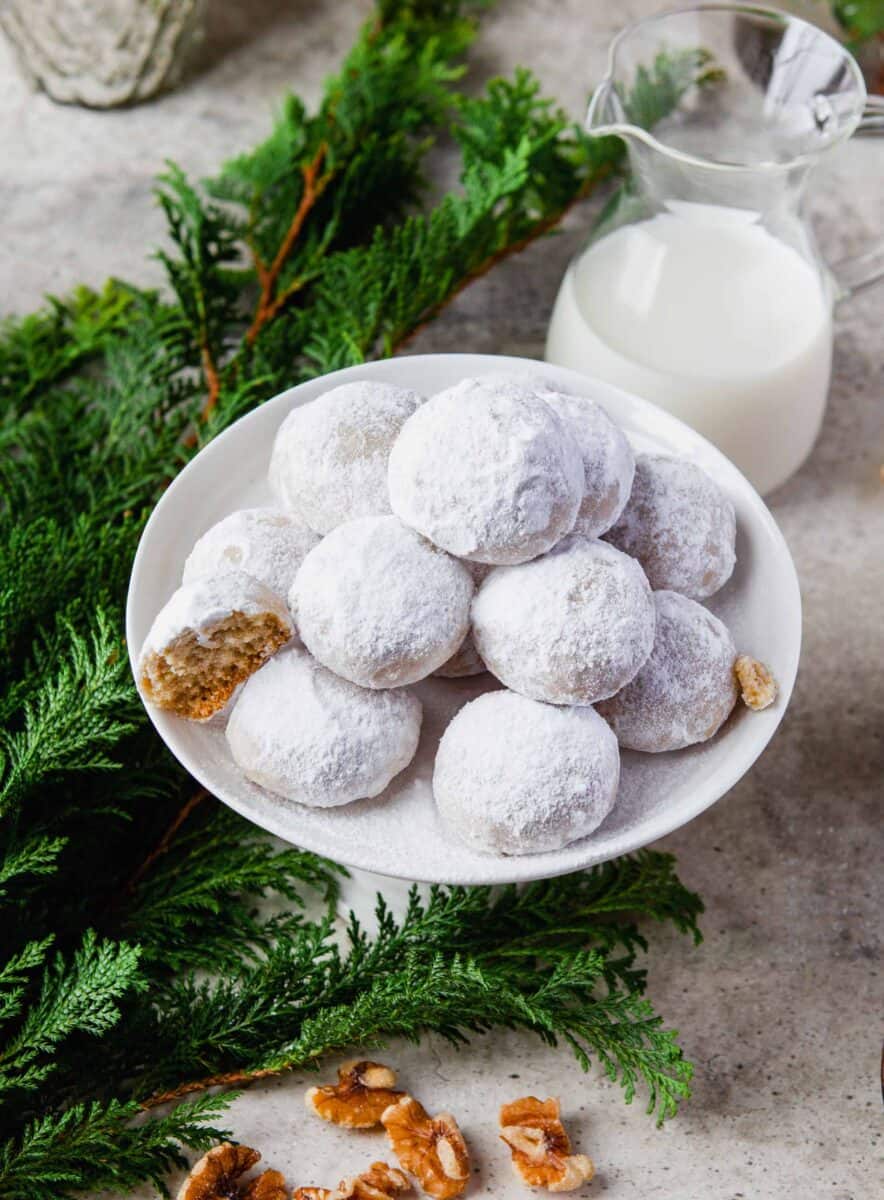 a pile of snowball cookies on a white plate with evergreen tree leaves next to it and a carafe of milk