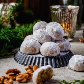 snowball cookies stacked on top of an upside down dark grey tart pan. whole walnuts and evergreen pine leaves can be seen in the scenery surrounding the snowball cookies. a dim candle is lit in the background as well