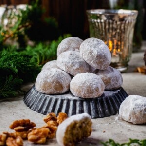 snowball cookies stacked on top of an upside down dark grey tart pan. whole walnuts and evergreen pine leaves can be seen in the scenery surrounding the snowball cookies. a dim candle is lit in the background as well