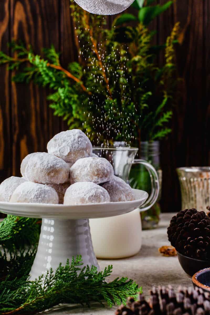 a stack of snowball cookies on a cake stand with powdered sugar being sprinkled above. pinecones in a bowl can be seen next to the cake stand