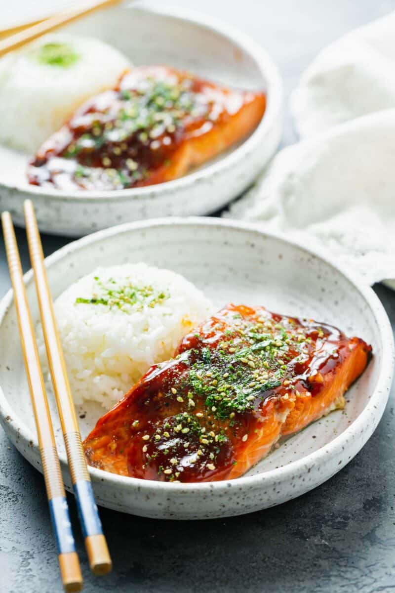 teriyaki glazed salmon on a speckled ceramic plate with furikake sprinkled on top next to a round mound of white rice with wooden chopsticks