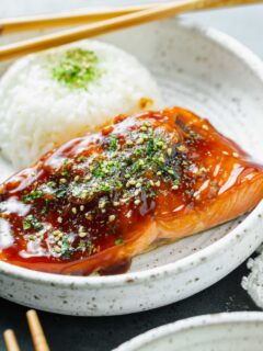 close up image of teriyaki glazed salmon on a speckled ceramic plate with furikake sprinkled on top next to a round mound of white rice with wooden chopsticks