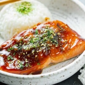 close up image of teriyaki glazed salmon on a speckled ceramic plate with furikake sprinkled on top next to a round mound of white rice with wooden chopsticks