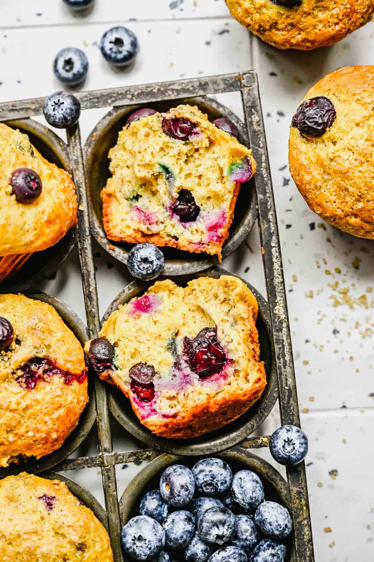 A banana blueberry muffins sliced in half next to whole muffins and fresh blueberries.