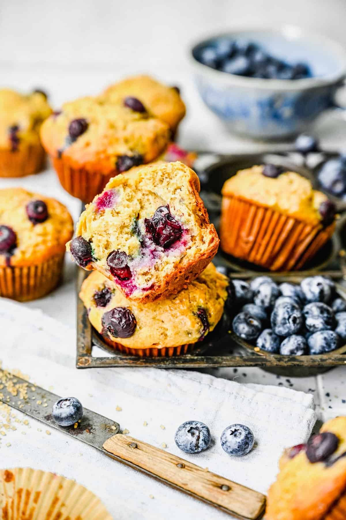 A banana blueberry muffins sliced in half next to whole muffins, a knife, and fresh blueberries.