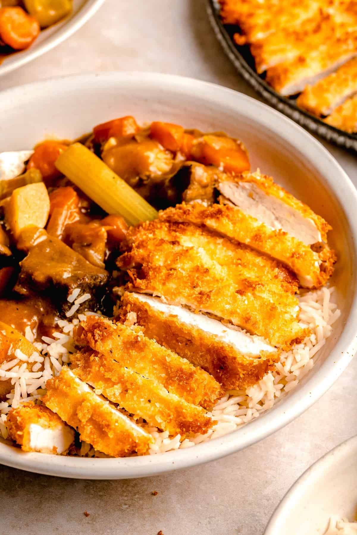 Katsu chicken curry served in a bowl over rice near a plate of chicken katsu and a bowl of curry.