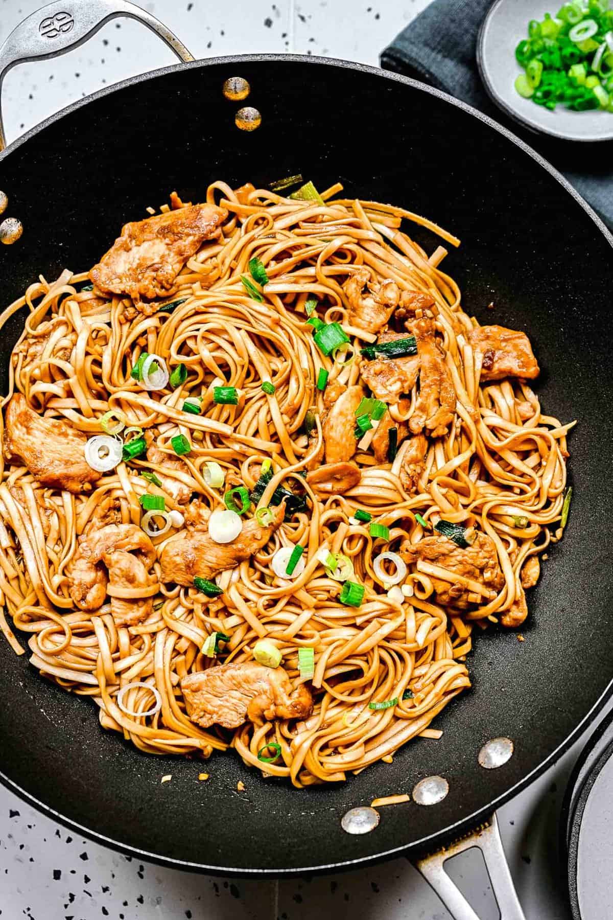 Chicken lo mein in a pan next to sliced scallions.