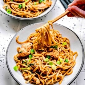 2 bowls of chicken lo mein with chopsticks next to sliced scallions.