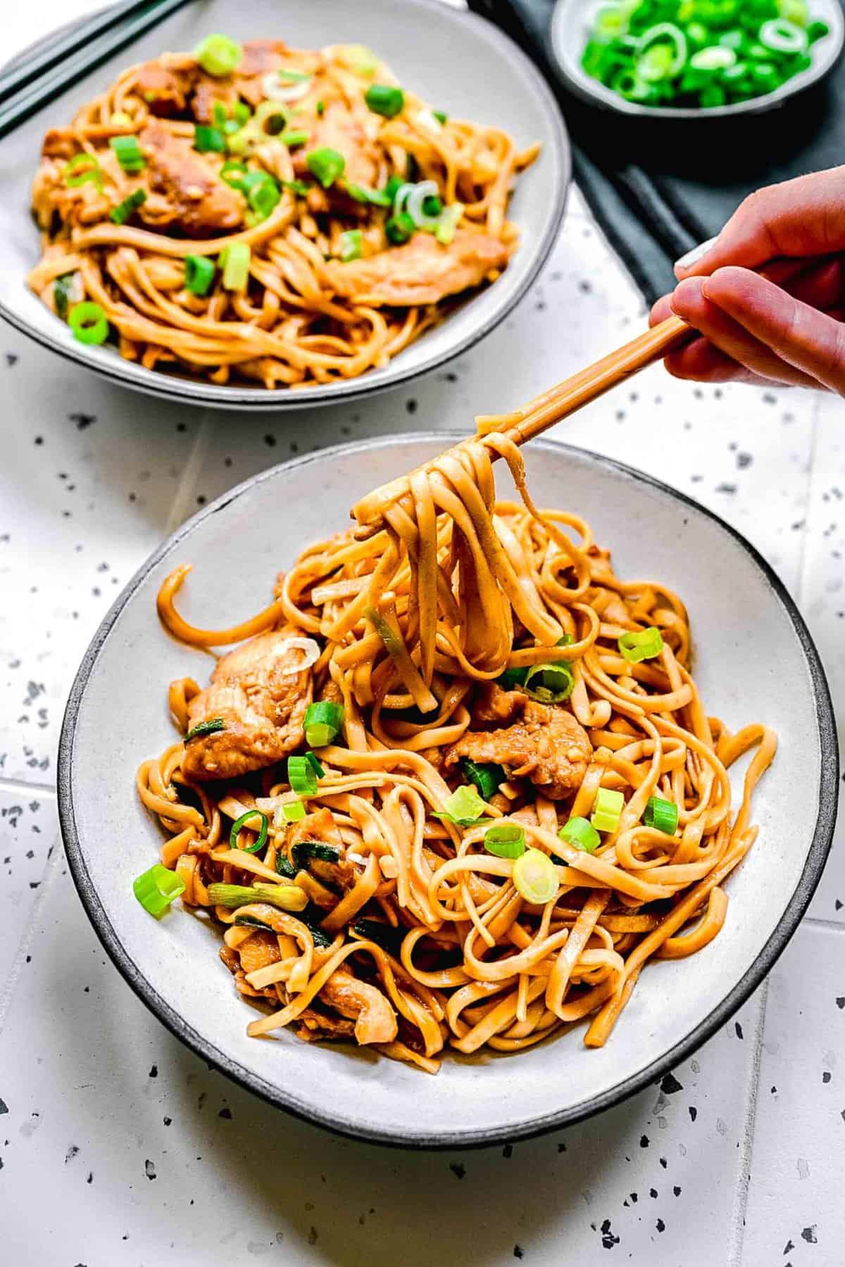 2 bowls of chicken lo mein with chopsticks next to sliced scallions.