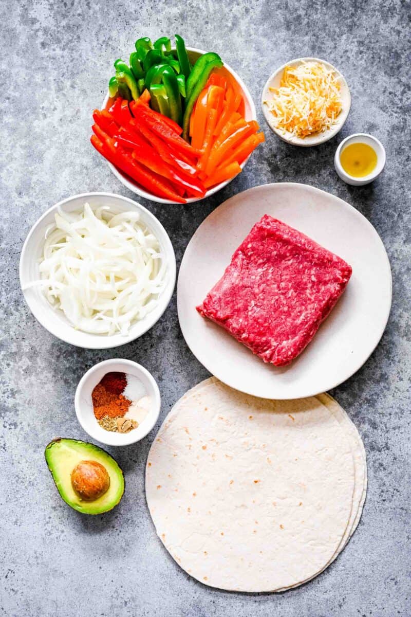 ingredients for ground beef fajitas: various colored bell peppers, shredded cheese, oil, ground beef, tortillas, avocado, seasonings, and sliced onions