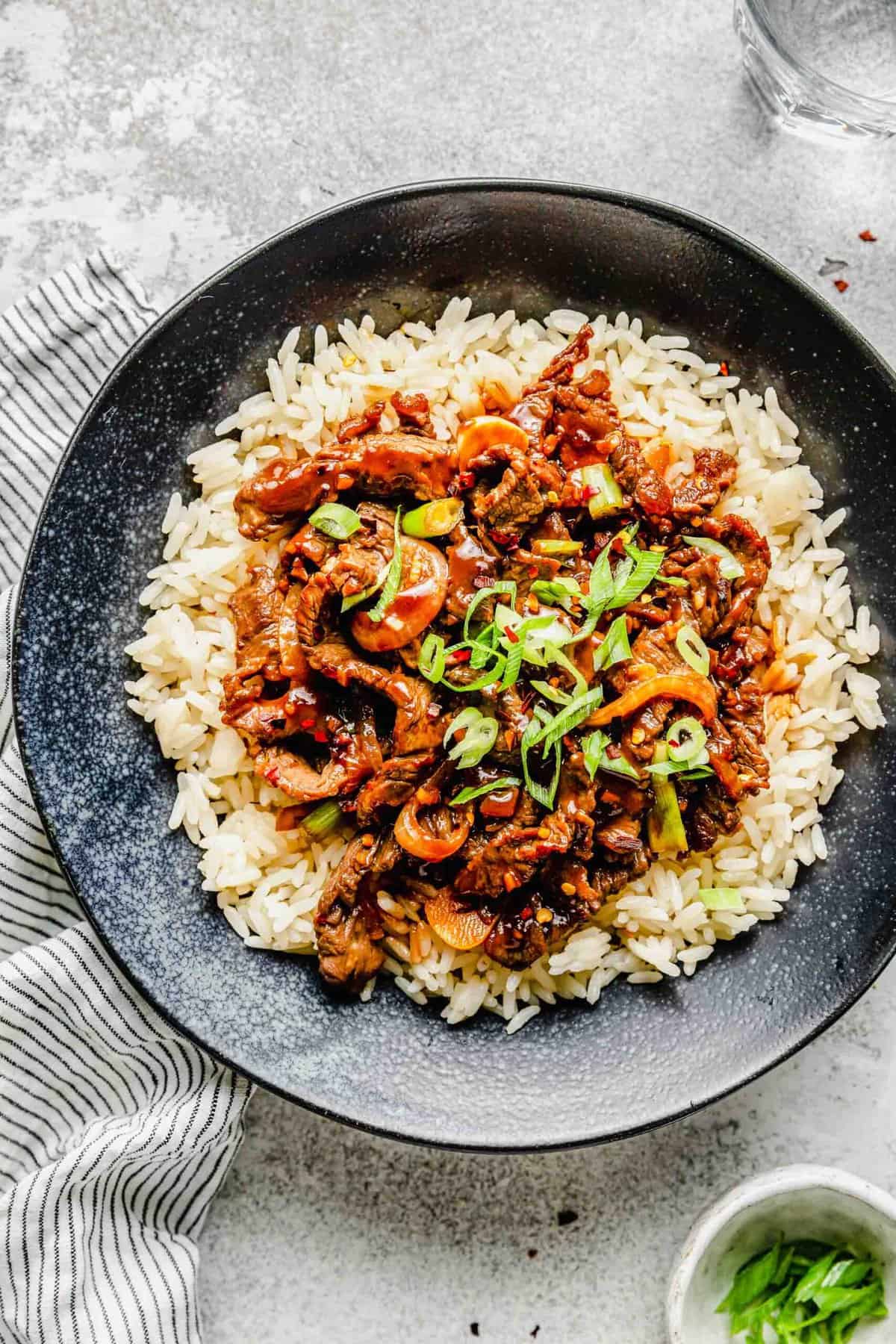 Korean beef bulgogi served over rice in a bowl next to a bowl of scallions.